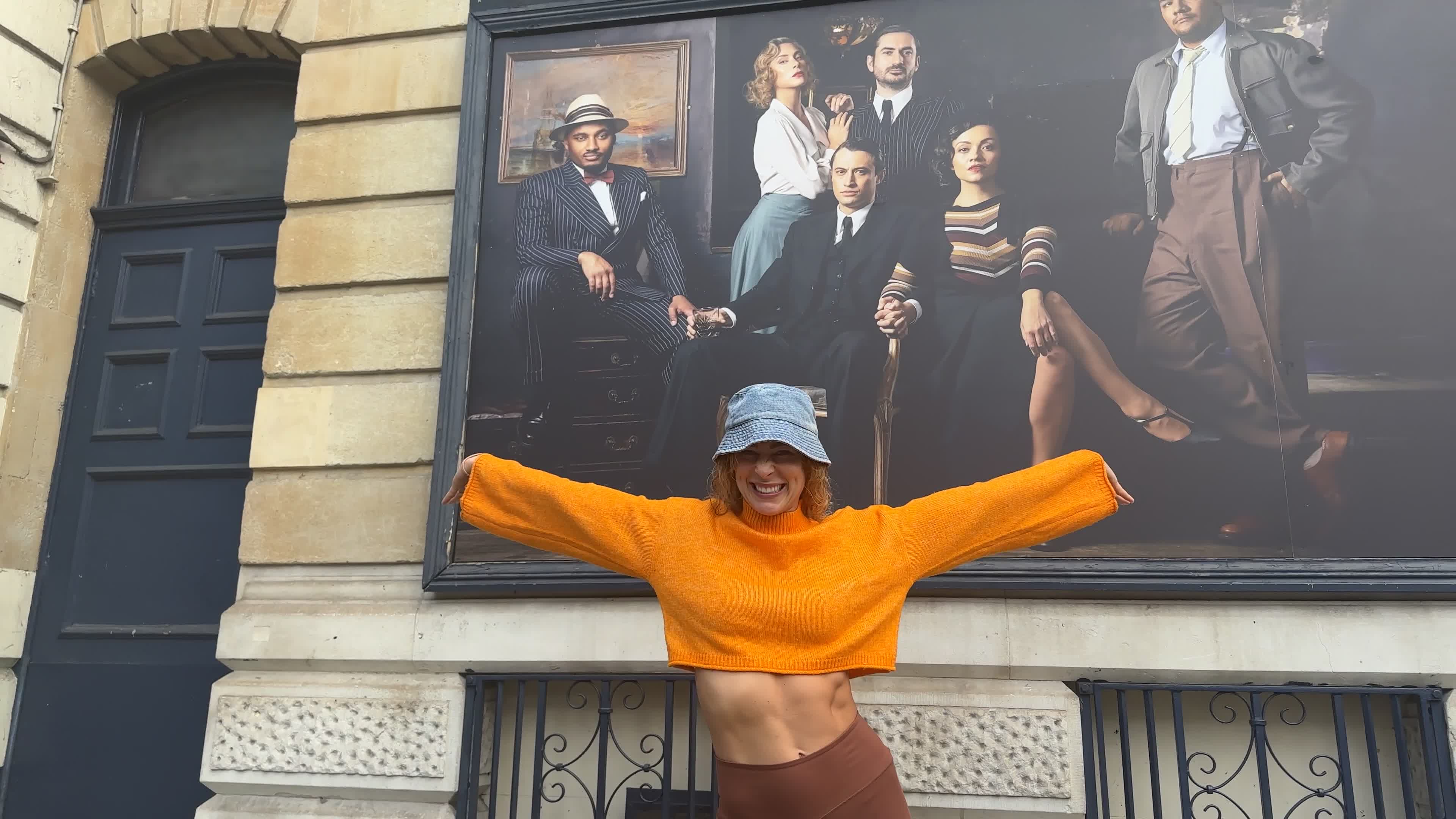 Jodie Steele outside the theatre, smiling with her arms outstretched in an orange cropped jumper